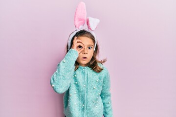 Little beautiful girl wearing cute easter bunny ears doing ok gesture shocked with surprised face, eye looking through fingers. unbelieving expression.