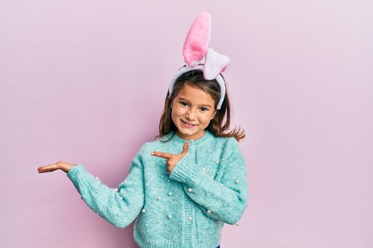 Little beautiful girl wearing cute easter bunny ears amazed and smiling to the camera while presenting with hand and pointing with finger.