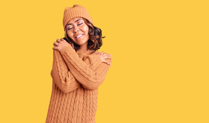 Young beautiful mixed race woman wearing wool sweater and winter hat hugging oneself happy and positive, smiling confident. self love and self care