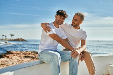 Young gay couple smiling happy sitting on the bench at the beach promenade.