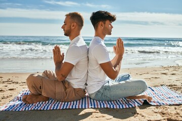Young gay couple doing yoga sitting at the beach.