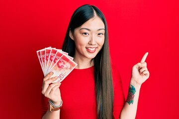 Young chinese woman holding 20 israel shekels banknotes smiling happy pointing with hand and finger to the side