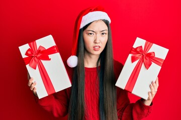 Young chinese woman wearing christmas hat and holding gifts clueless and confused expression. doubt concept.