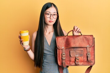 Young chinese woman holding leather bag and drinking a take away coffee relaxed with serious expression on face. simple and natural looking at the camera.