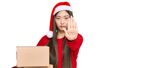 Young chinese woman wearing christmas hat holding delivery package with open hand doing stop sign with serious and confident expression, defense gesture