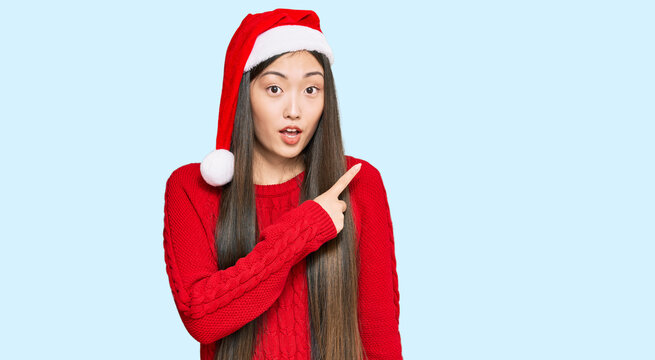 Young chinese woman wearing christmas hat surprised pointing with finger to the side, open mouth amazed expression.