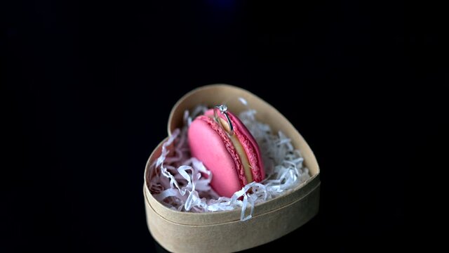 pink macaroon cookies in a heart-shaped box with a wedding ring on a black table