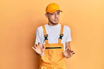 Hispanic young man wearing handyman uniform disgusted expression, displeased and fearful doing disgust face because aversion reaction.