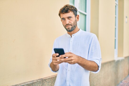 Handsome man with beard wearing casual white shirt on a sunny day smiling happy outdoors using smartphone