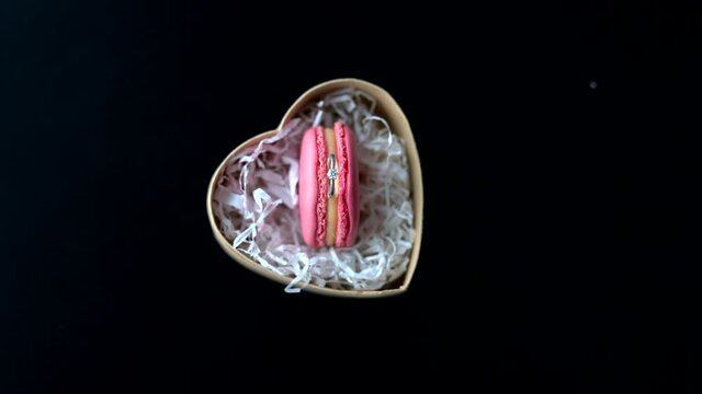 pink macaroon cookies in a heart-shaped box with a wedding ring on a black table
