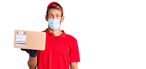 Handsome blond man with beard holding delivery box wearing medical mask scared and amazed with open mouth for surprise, disbelief face