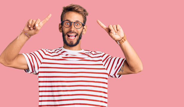 Handsome blond man with beard wearing casual clothes and glasses smiling amazed and surprised and pointing up with fingers and raised arms.