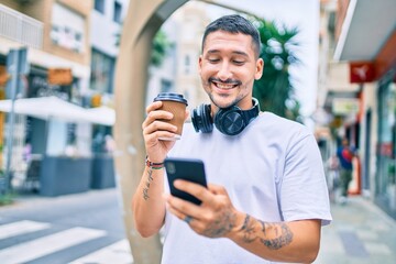 Young hispanic man using smartphone and headphones drinking coffee at street of city