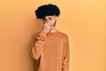 Fototapeta na wymiar Young african american man with afro hair wearing casual winter sweater smelling something stinky and disgusting, intolerable smell, holding breath with fingers on nose. bad smell