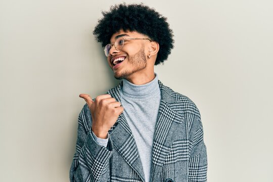 Young african american man with afro hair wearing casual clothes  glasses pointing thumb up to the side smiling happy with open mouth