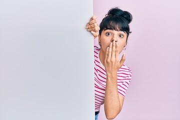 Young hispanic girl holding blank empty banner covering mouth with hand, shocked and afraid for mistake. surprised expression