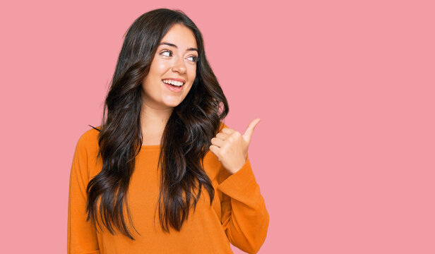 Beautiful brunette young woman wearing casual orange sweater smiling with happy face looking and pointing to the side with thumb up.
