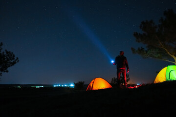 Man with bright flashlight near camping tents outdoors at night - obrazy, fototapety, plakaty