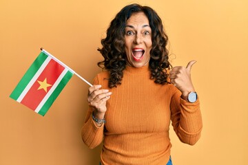 Middle age hispanic woman holding suriname flag pointing thumb up to the side smiling happy with...