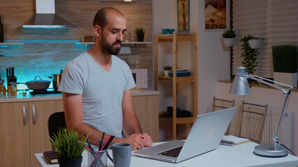Businessman waving during video conference while working from home at night. Employee using modern...