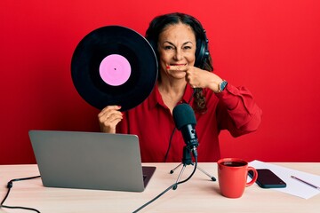 Beautiful middle age woman working at radio studio holding vinyl disc smiling happy pointing with hand and finger