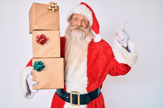 Old senior man with grey hair and long beard wearing santa claus costume holding presents surprised with an idea or question pointing finger with happy face, number one