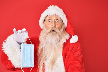 Old senior man wearing santa claus costume holding safety mask scared and amazed with open mouth for surprise, disbelief face