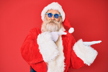 Old senior man wearing santa claus costume and sunglasses amazed and smiling to the camera while presenting with hand and pointing with finger.
