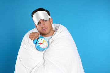 Man in sleeping mask wrapped with blanket holding alarm clock on blue background