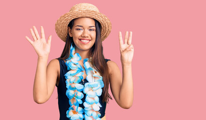 Young beautiful latin girl wearing hawaiian lei and summer hat showing and pointing up with fingers number eight while smiling confident and happy.
