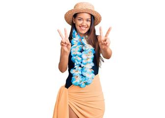 Obraz na płótnie Canvas Young beautiful latin girl wearing hawaiian lei and summer hat smiling looking to the camera showing fingers doing victory sign. number two.