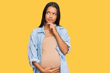 Beautiful hispanic woman expecting a baby showing pregnant belly serious face thinking about...