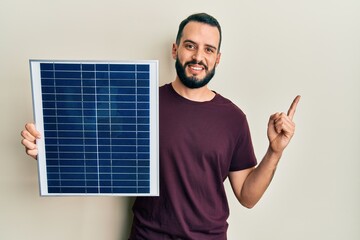 Young man with beard holding photovoltaic solar panel smiling happy pointing with hand and finger...