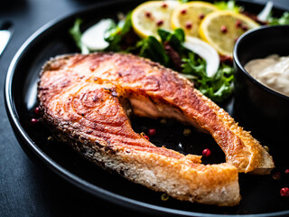 Fried salmon steak, mayonnaise and mix of vegetables served on black plate on wooden table
