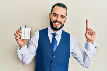 Young man with beard drinking whiskey from flask surprised with an idea or question pointing finger with happy face, number one