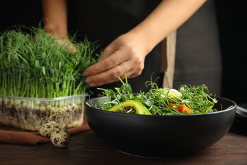 Woman picking fresh organic microgreen at wooden table, focus on bowl of delicious salad