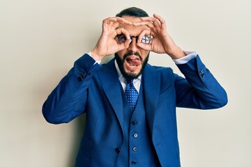 Young man with beard wearing business suit and tie doing ok gesture like binoculars sticking tongue out, eyes looking through fingers. crazy expression.