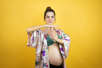 Young beautiful woman pregnant expecting baby wearing pajama over isolated yellow background Doing time out gesture with hands, frustrated and serious face