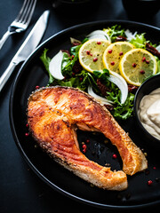 Fried salmon steak, mayonnaise and mix of vegetables served on black plate on wooden table
