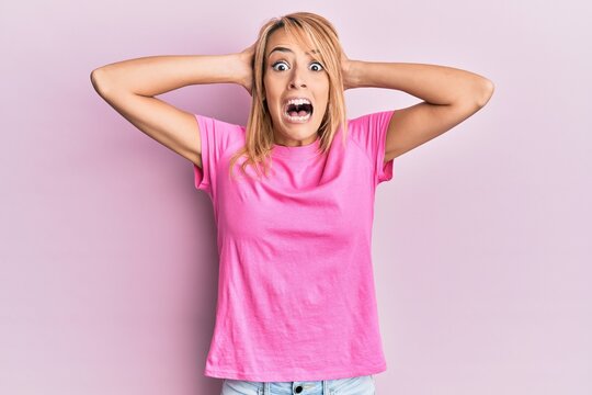 Beautiful blonde woman wearing casual pink tshirt crazy and scared with hands on head, afraid and surprised of shock with open mouth