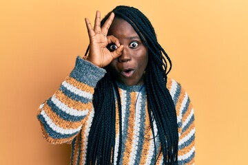 Young black woman with braids wearing casual winter sweater doing ok gesture shocked with surprised face, eye looking through fingers. unbelieving expression.