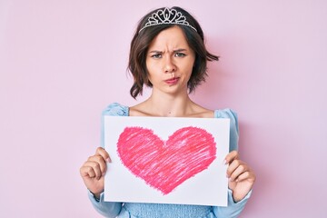 Young beautiful girl wearing princess crown holding heart draw skeptic and nervous, frowning upset because of problem. negative person.