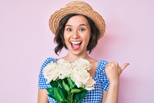 Young beautiful girl wearing summer hat holding flowers pointing thumb up to the side smiling happy with open mouth