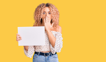 Young blonde woman with curly hair holding sale poster covering mouth with hand, shocked and afraid...