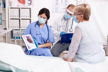 Nurse explaining heart diagnosis in cardiology to senior woman on tablet pc in time of covid pandemic. Medical assistant having a conversation with elder patient during coronavirus outbreak.