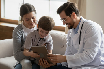 Caring male doctor in white uniform use tablet consult small boy patient at consultation in hospital with mom. Man pediatrician and little child with mother look at pad screen at visit in clinic. - Powered by Adobe