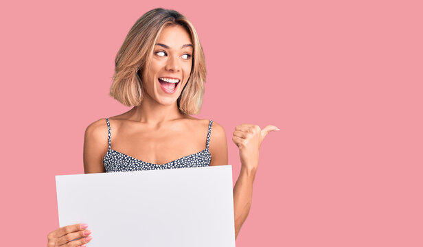 Beautiful caucasian woman holding blank empty banner pointing thumb up to the side smiling happy with open mouth