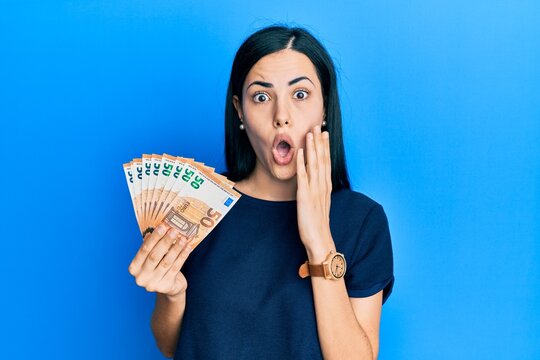 Beautiful young woman holding bunch of 50 euro banknotes afraid and shocked, surprise and amazed expression with hands on face