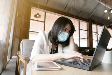 Asian businesswoman is wearing mask prevent for civid-19 and online working with laptop in office. She is searching social on internet at coffee shop. Healthy and business with techonology concept