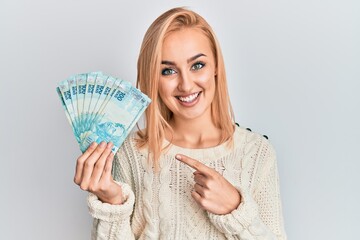 Beautiful caucasian blonde woman holding 100 brazilian real banknotes smiling happy pointing with hand and finger